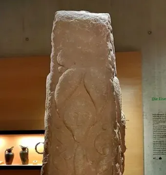 Pfalzfeld Obelisk, a weathered carved stone column displayed on an orange platform in a museum.