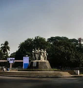 Raju Memorial Sculpture stands on a circular tiered base at a road junction, surrounded by trees, banners, and people nearby.