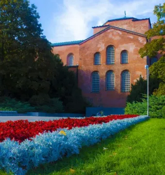 Unknown Soldier Monument beside a reddish-brick building with arched windows, framed by trees and flower beds.