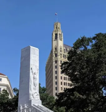 The Alamo Cenotaph stands in front of the Morgan Hotel tower under a clear blue sky.