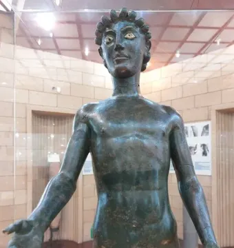 Efebo de Selinunte bronze statue of a nude young man with outstretched arms and green patina behind glass