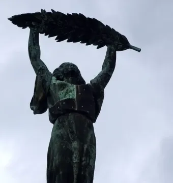 Liberty Statue bronze figure raising a leafy branch against a cloudy sky.