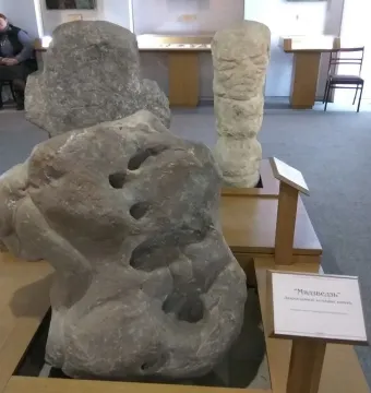 Škloŭ Idol stone sculpture with a rough grey surface and rounded indentations, displayed on a wooden stand.