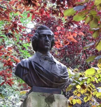 Dark metallic bust of Frédéric Chopin mounted on a weathered stone block amid red and green foliage.