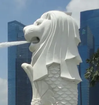 The Merlion statue with water jetting from its mouth, set against modern city buildings and a blue sky.
