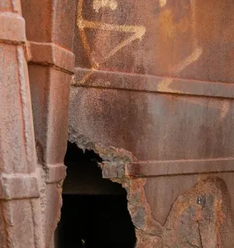 Close-up of the Monument to the Azoreans showing a heavily rusted metal surface with faint graffiti and a jagged hole near the bottom.