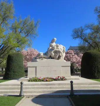 White stone war memorial with sculptural figures on a tiered base, surrounded by lawns and flowering trees.