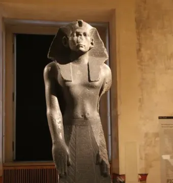 Amenemhat III statue of a standing bare-chested male figure wearing a striped headdress and pleated kilt indoors.