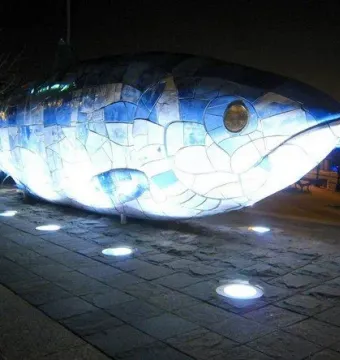 The Big Fish sculpture covered in blue and white mosaic tiles, lit up at night on a waterfront plaza.