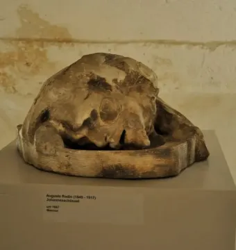 Weathered stone sculpture of the Head of Saint John the Baptist displayed on a pedestal in a museum setting.