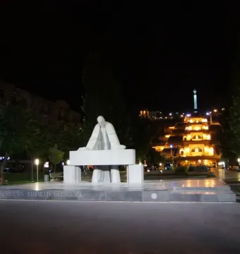 Monument to Alexander Tamanyan in a lit plaza at night, with a grand illuminated building complex and distant spire behind it.
