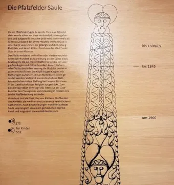 Illustrated Pfalzfeld Obelisk shown in black outline with decorative faces and patterns on a light wooden background.