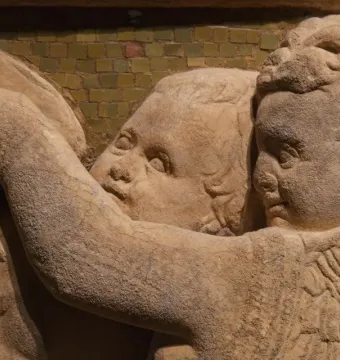 Close-up of a marble relief from the Pulpit of Prato Cathedral showing winged figures, faces, and a hand in front of a mosaic background.