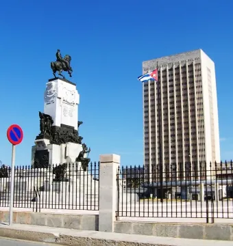 Antonio Maceo Monument with an equestrian statue on a tall white pedestal, a high-rise building and Cuban flag behind it.
