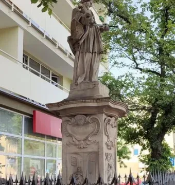 Weathered stone statue of John of Nepomuk on a sculpted pedestal behind a decorative metal fence.