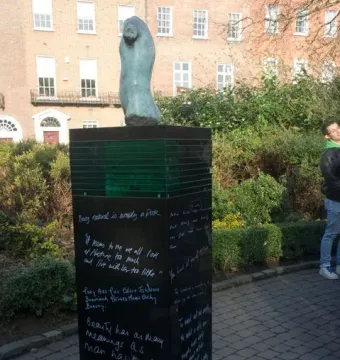 Oscar Wilde Memorial Sculpture on a tall dark pillar with white inscriptions beside a paved path and greenery.