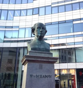 Schadow Monument bust on a tall stone base with a modern curved building behind it.