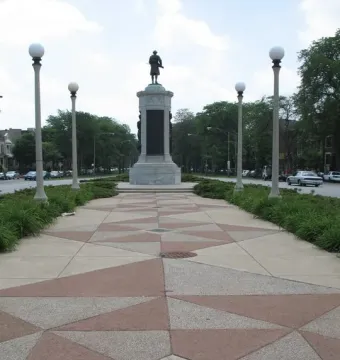 Victory Monument rising at the end of a patterned pedestrian walkway with lampposts and trees nearby.