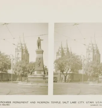 Brigham Young Monument with a standing statue atop a tall pedestal in a tree-lined street median, with a large multi-spired building in the background.