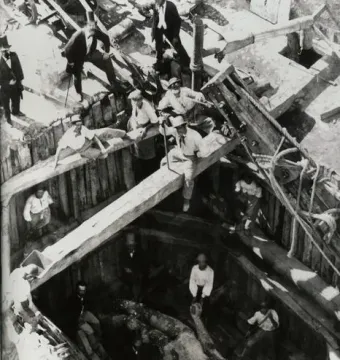 A large reclining Hercules statue is being unearthed from a timber-lined excavation pit as several men work around it.