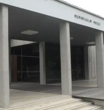Front entrance of the Memorial Museum Jasenovac with grey concrete pillars, glass windows, and steps leading up to the door.
