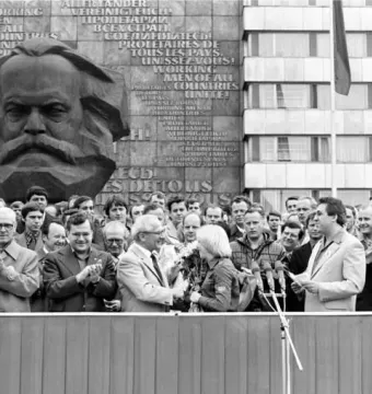 Karl Marx Monument beside a crowd gathered for an outdoor event, with a building facade behind it bearing a multilingual slogan.