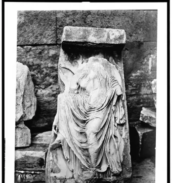 Fragmented classical relief of Nike untying her sandal, showing a draped headless female figure with one hand reaching toward a raised leg.