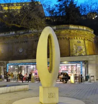 Zero Kilometre Stone on a paved plaza with a cafe and illuminated brick structure in the background.