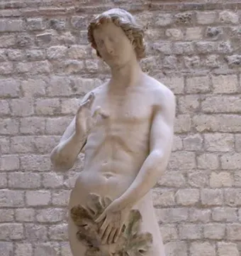 Stone statue of Adam standing nude with a leaf covering his groin and one hand raised.