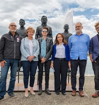 Seven people pose beside the Anything to Say? sculpture with three seated bronze figures and an empty chair by the water in Geneva.
