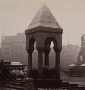 Bagley Memorial Fountain in a hazy city square, with a pyramidal stone top, arched openings, and ornate carvings.