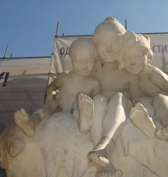 Light-colored stone sculpture of three youths on a rocky base beneath a bright blue sky, with scaffolding and a Cyrillic banner behind them.