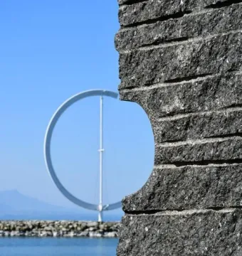 The Éole ring sculpture is seen through an opening in a dark stone wall, with water and sky behind it.