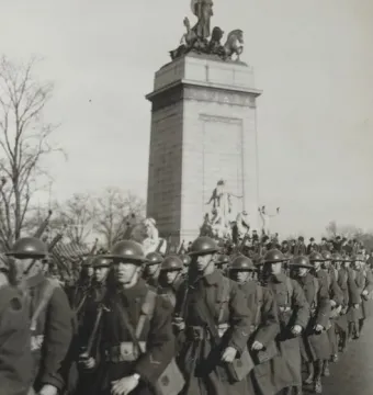 USS Maine National Monument with a sculptural figure and horses atop a tall stone base, seen behind marching soldiers.