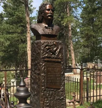 Bronze bust of Wild Bill Hickok on a textured pedestal with a pistol and hat relief, set behind a fence among pine trees.