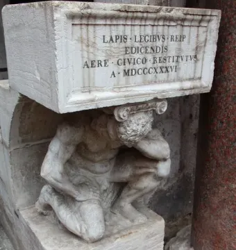Il Gobbo di Rialto, a weathered stone figure crouching beneath an inscribed block beside a reddish column.
