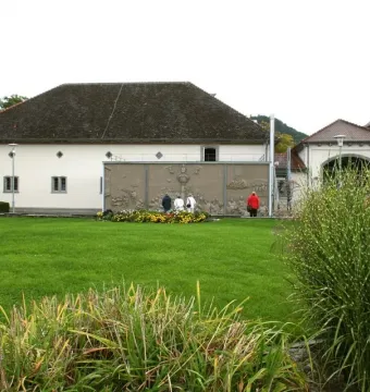 The stone relief Ludwigs Erbe mounted on an exterior wall, with four people standing in front of it on a lawn.
