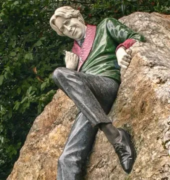 Oscar Wilde Memorial Sculpture showing a reclining man beside a rough boulder with leafy trees behind him.