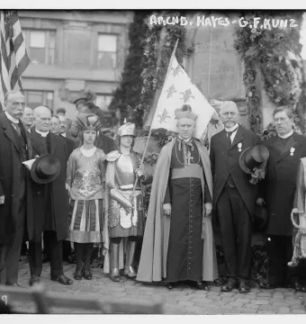 A dark Joan of Arc monument draped with garlands and foliage stands behind a cardinal and children in historical costumes.