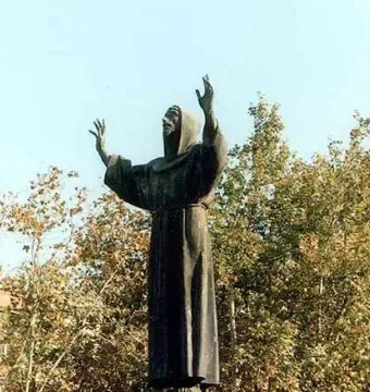 Monument to Saint Francis of Assisi, a dark bronze hooded figure with raised hands against a clear sky and autumn trees.