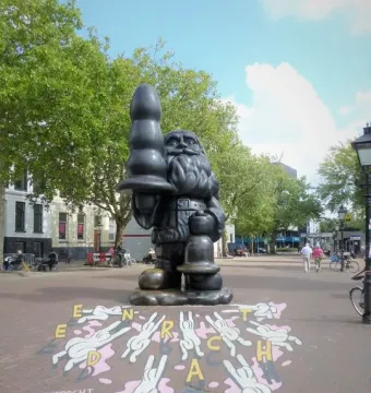 Dark Santa Claus sculpture standing in a paved square and holding a large black phallic-shaped object.