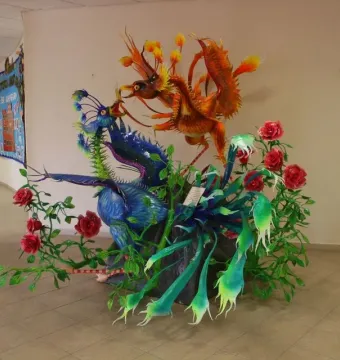 Alebrije sculpture with a blue-purple winged creature beside an orange-red bird-like figure, surrounded by green foliage and red roses.