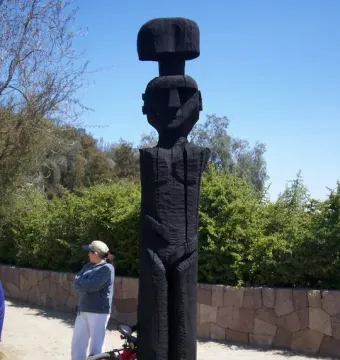 Chemamull wooden sculpture of a stylized human figure beside a bicycle on a dirt path.