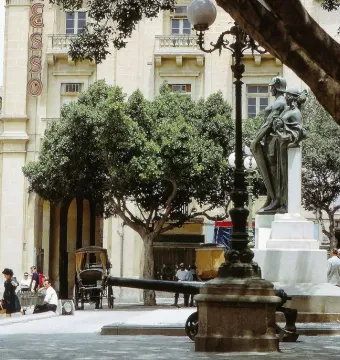 Great Siege Monument with three bronze draped figures on a white base beside a street lamp and cannon.