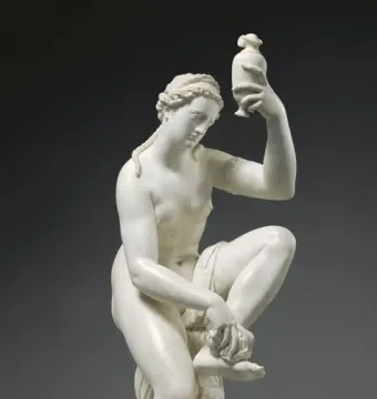 White sculpture of a kneeling female figure holding a small vessel aloft beside a draped pillar.