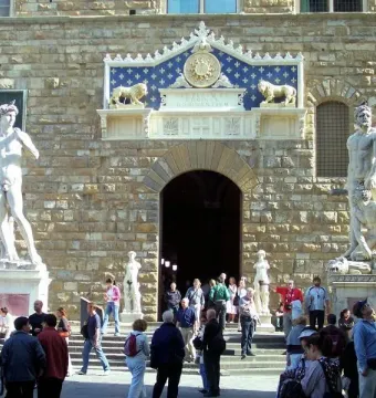 Hercules and Cacus marble sculpture beside a stone building entrance, with two figures on an ornate pedestal surrounded by onlookers.