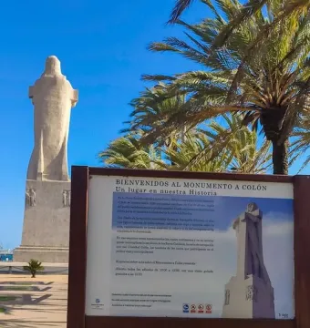 Information sign for the Monument to Columbus with the statue in the background against a blue sky.