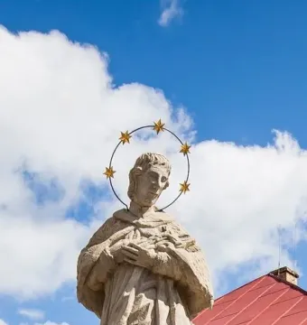 Statue of St. John of Nepomuk viewed from below against a blue sky, wearing a halo of stars and holding an object to his chest.