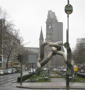 Light-grey tubular sculpture by Brigitte und Martin Matschinsky-Denninghoff in a city plaza, with a historic church ruin in the background.