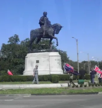 Gen. Beauregard equestrian statue on a tall white pedestal in a grassy traffic circle, with several people nearby displaying Confederate battle flags.
